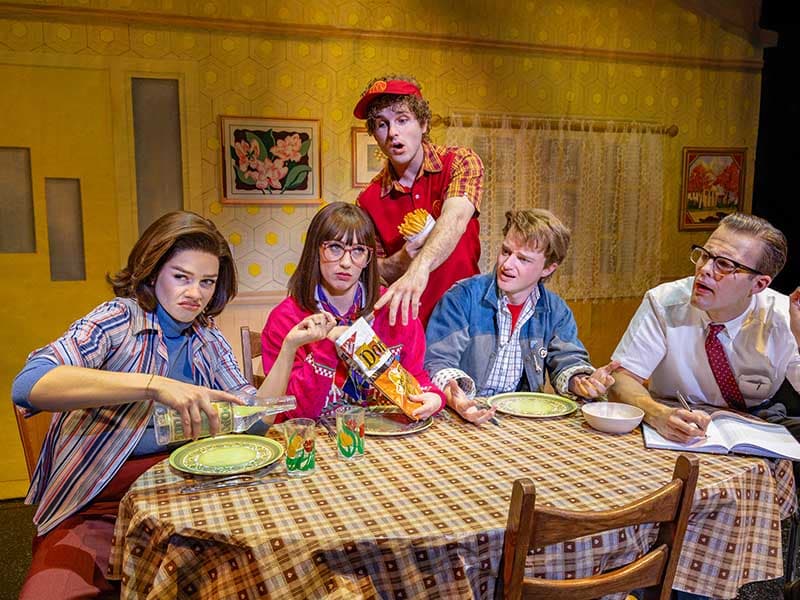 The McFly family gathers for an unconventional dinner scene in the touring production of Back to the Future: The Musical. Lucas Hallauer as Marty McFly sits alongside his eccentric relatives, surrounded by 1980s-inspired set design and nostalgic props.