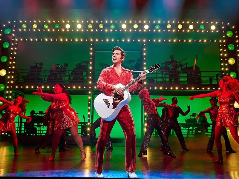 Nick Fradiani as Neil Diamond holds a white acoustic guitar while wearing a red sequin outfit. Dancers appear on stage behind him.