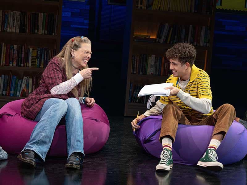 Kimberly and Seth sit on beanbags facing each other. They are both laughing, and Seth holds a notebook.